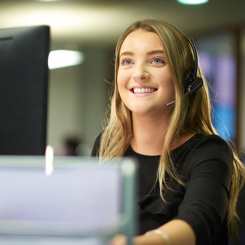 a teenage office worker sits at her desk and speaks on her telephone headset . She is smiling and looks happy to be working in a big open plan office .